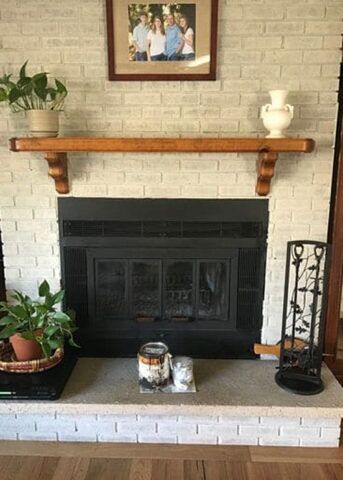 Light gray brick fireplace with a wooden mantel holding a framed family photo, a potted plant, and a white vase. Below is a black metal fireplace insert, candles on the hearth, and a black wrought iron fireplace tool holder on the right.